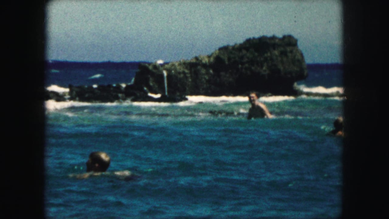1959 8mm frame showing swimmers in blue ocean water with dark rocks and white surf in the background.