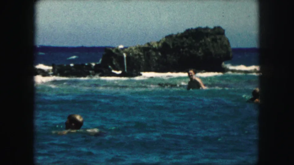 1959 8mm frame showing swimmers in blue ocean water with dark rocks and white surf in the background.