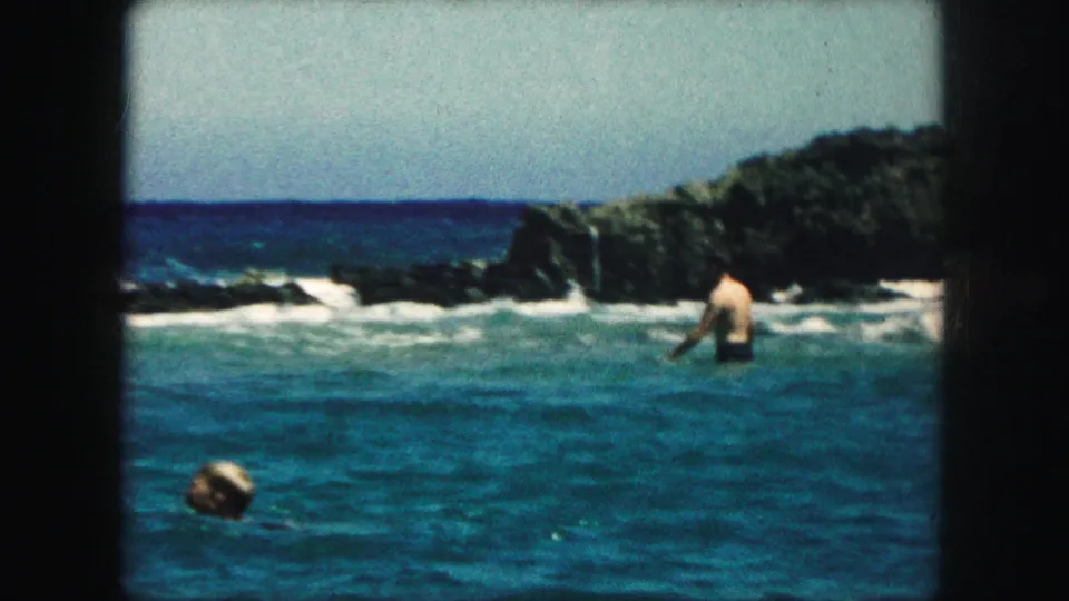 1959 8mm home-movie frame of a swimmer standing near surf with another swimmer partly visible in the foreground.