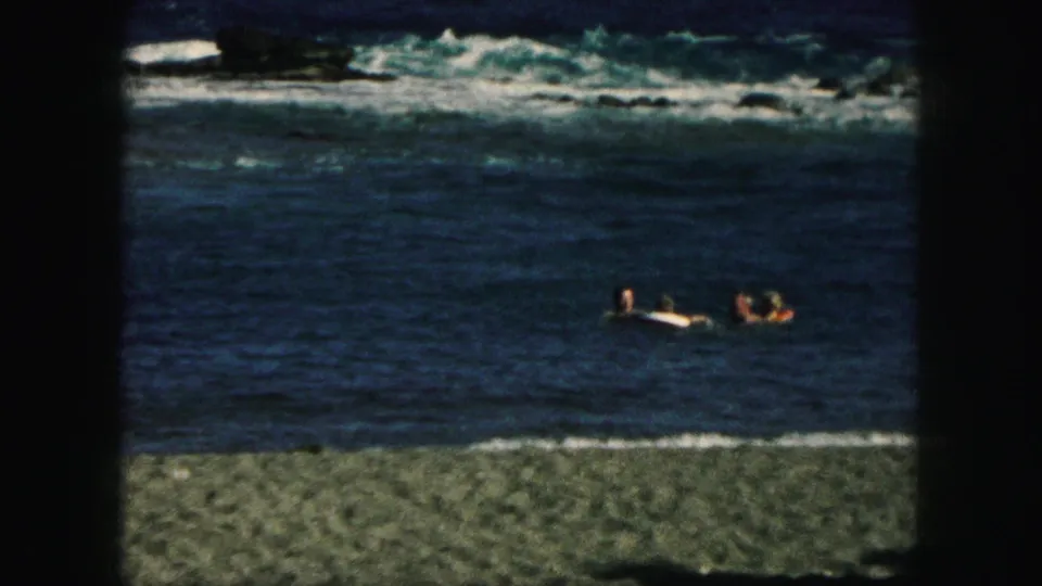 1959 8mm frame with swimmers offshore, a rocky or pebbly beach in front, and waves breaking in the distance.
