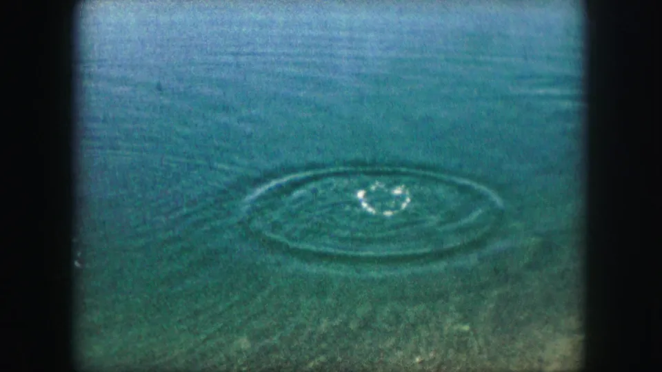 1959 8mm frame of green-blue pond water in the Kansas City area, with circular ripples expanding after a splash.