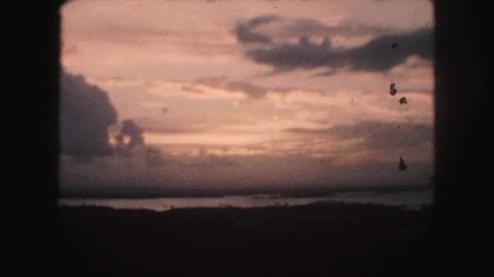 1959 8mm sunset frame over a Kansas City area pond or lake, with warm clouds, a low horizon, and visible film dust.