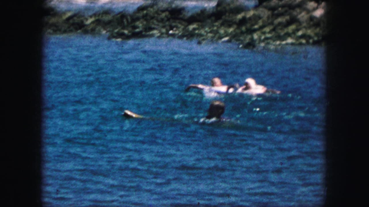 1959 8mm home-movie frame from Kansas City metadata showing people swimming in blue lake water near a rocky shoreline.