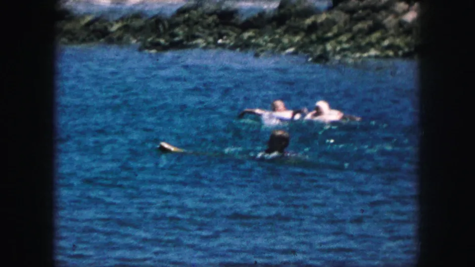 1959 8mm home-movie frame from Kansas City metadata showing people swimming in blue lake water near a rocky shoreline.