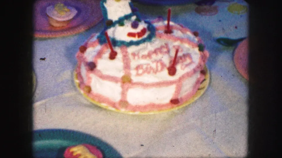 Close 8mm frame of a frosted birthday cake with red candles and colorful decoration on a party table, from the Kansas City 1959 clip metadata.