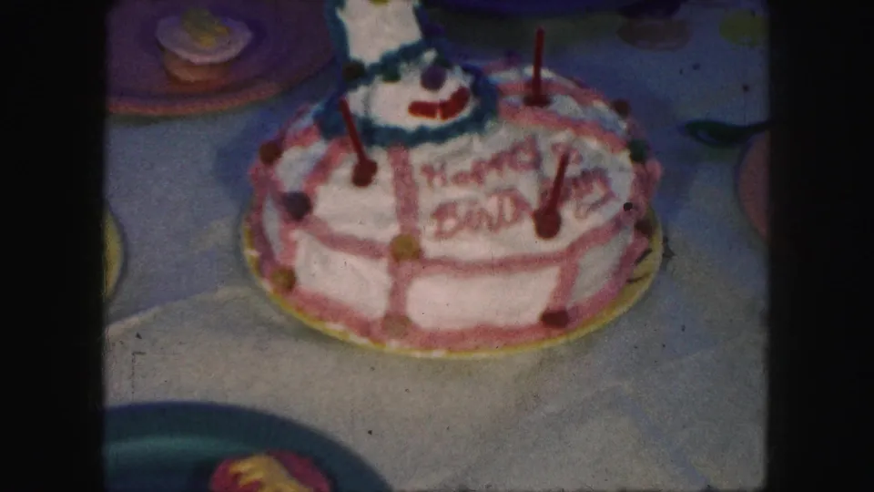 Dim, soft-focus 1959 home-movie close-up of the decorated birthday cake, with candles and nearby paper plates visible on the table.