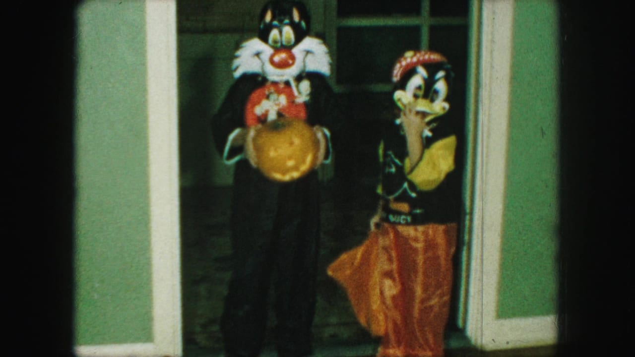 In 1959 Kansas City home-movie footage, two children in cartoon-style Halloween costumes pose in a doorway with a carved pumpkin.