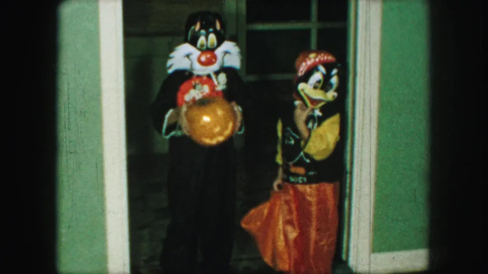 A child dressed as a cat holds a carved pumpkin while another child in a duck-like costume poses beside the doorway.
