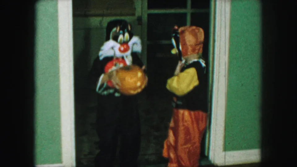 The child with the pumpkin faces forward as the duck-costumed child turns sideways near the doorway in the 1959 home movie.