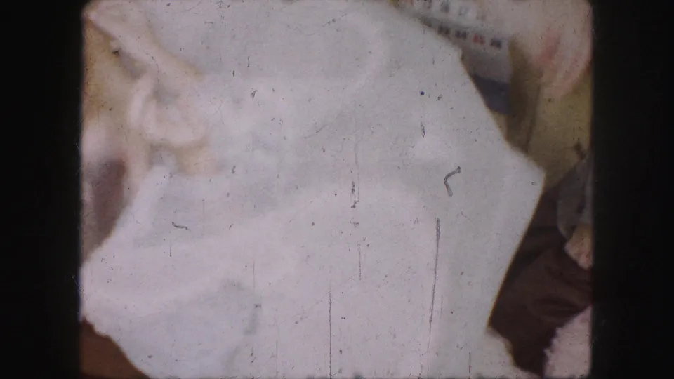 1959 Kansas City home-movie frame showing hands adjusting a white dress in a private interior, with a wall calendar behind.