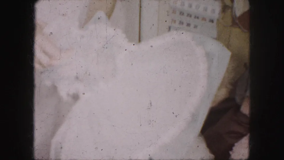 1959 Kansas City interior frame with a white dress held close to camera and a partially visible person near a wall calendar.