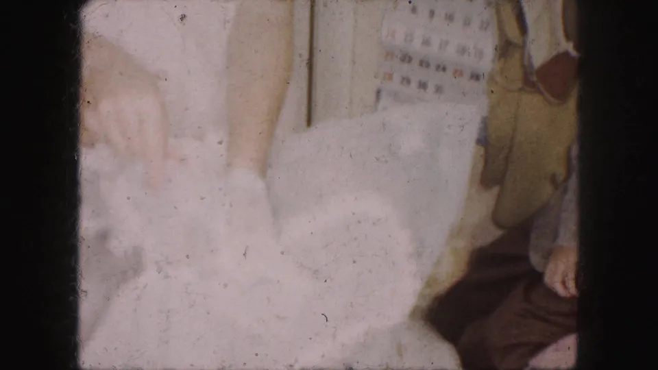 1959 Kansas City home-movie frame showing close hands at a white dress, with garments and a calendar on the room wall.