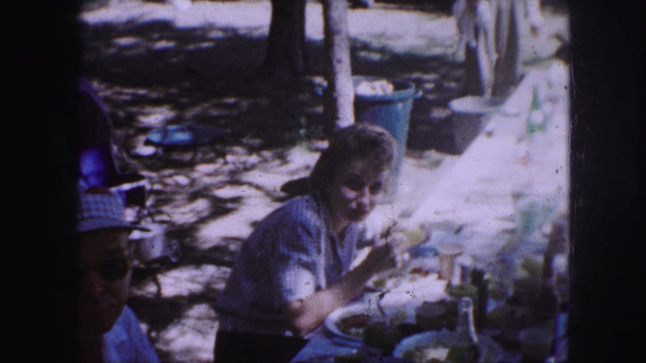 1959 Kansas City home-movie frame showing a woman at a park picnic table eating while picnic items and trees sit behind her.
