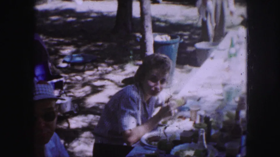 1959 Kansas City home-movie frame showing a woman at a park picnic table eating while picnic items and trees sit behind her.