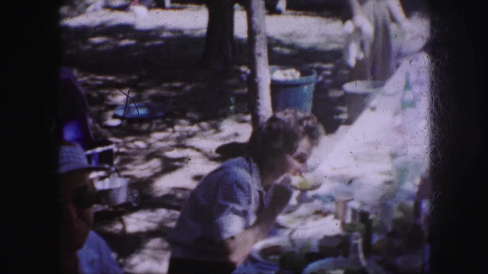A 1959 Kansas City picnic frame shows a woman seated outdoors, eating at a table with plates, cups, trees, and a blue trash can nearby.