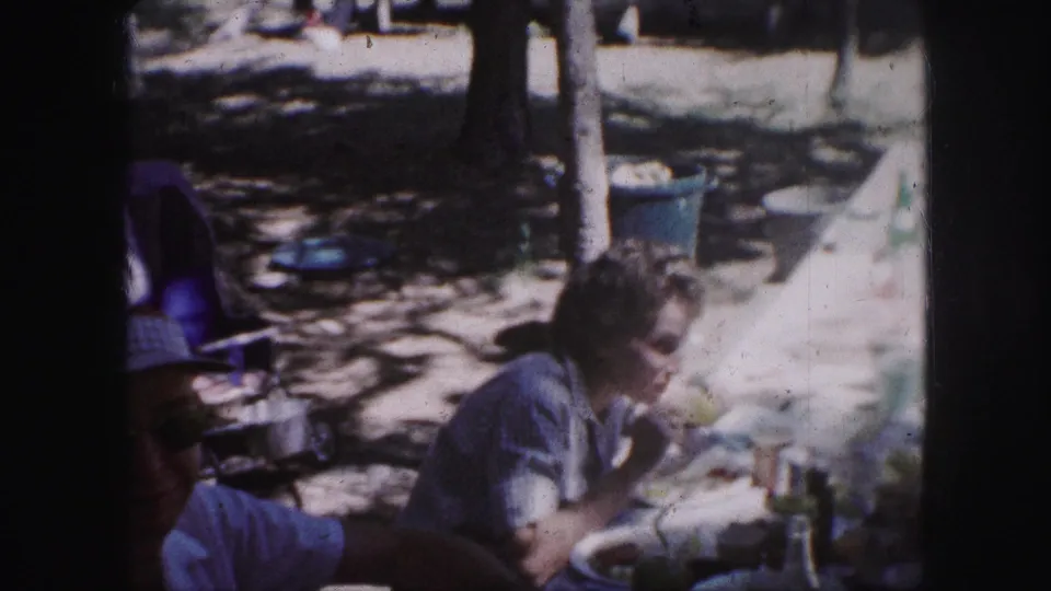Kansas City 1959 home-movie frame of a park picnic with a seated woman, partial adult figure, dishes, cups, and sun-dappled ground.