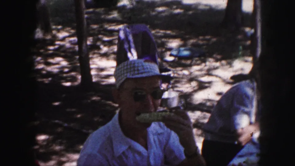 A 1959 Kansas City park picnic frame shows a man wearing sunglasses and a patterned cap eating corn on the cob, with another adult nearby.