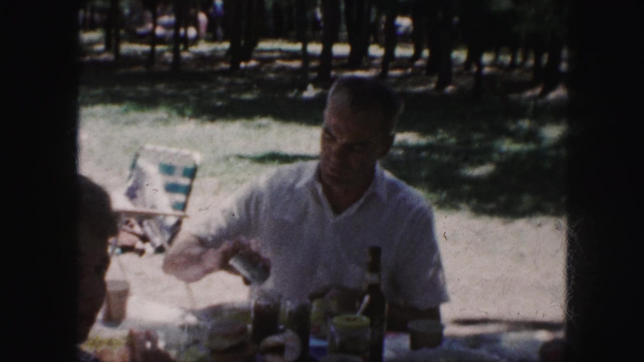 1959 Kansas City home-movie frame showing a man in a white short-sleeve shirt at a shaded picnic table with food and drink items.