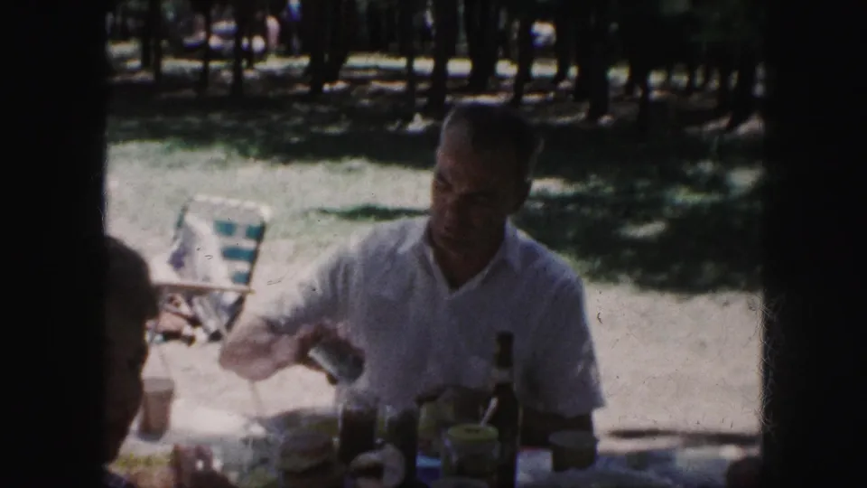 1959 Kansas City home-movie frame showing a man in a white short-sleeve shirt at a shaded picnic table with food and drink items.