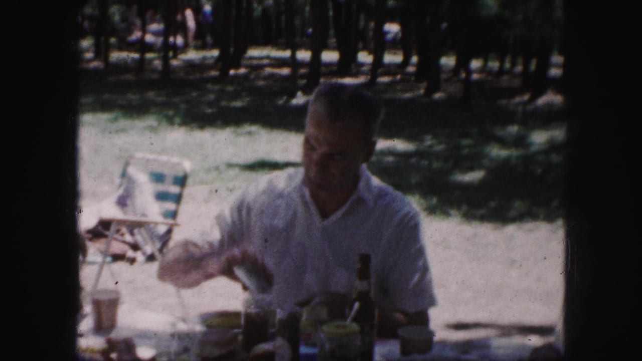 1959 Kansas City picnic frame with a man seated beneath trees, reaching at a table with cups, bottles, and picnic food visible.