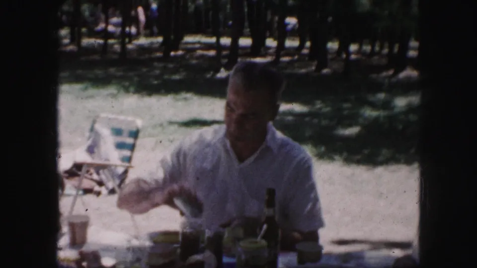 1959 Kansas City picnic frame with a man seated beneath trees, reaching at a table with cups, bottles, and picnic food visible.