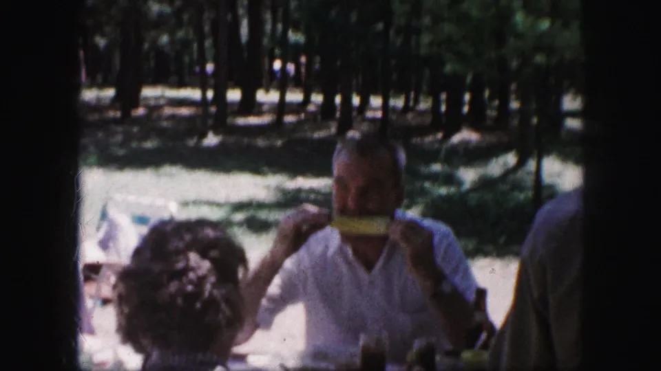 1959 Kansas City home-movie frame showing a man eating corn on the cob at a shaded picnic table, with other adults partly visible nearby.