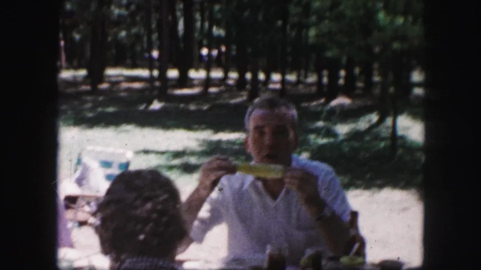 1959 Kansas City picnic frame with a man eating corn on the cob at a table under trees, seen through dark 8mm frame borders.