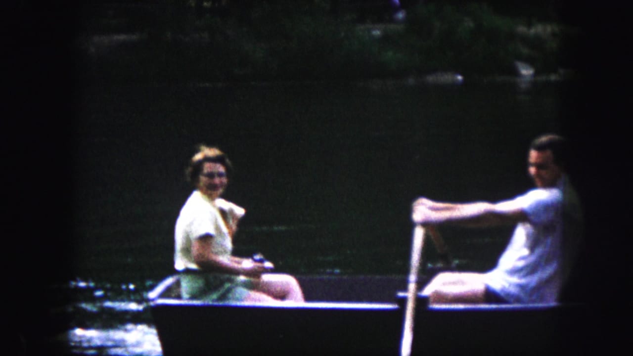 Woman holding a small dog faces a man rowing a dark rowboat on a lake.