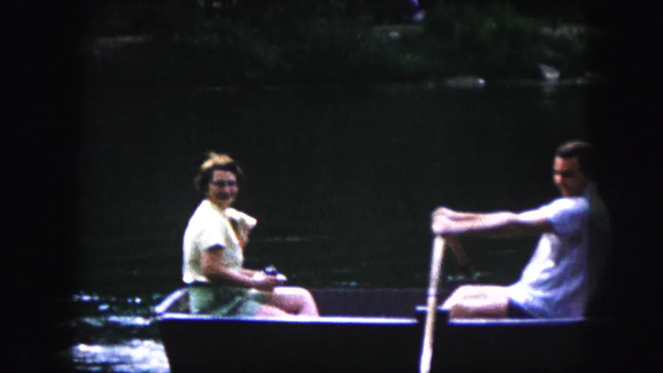 In 1959 Kansas City home-movie footage, a woman with a small dog sits opposite a man rowing a dark boat on calm water.