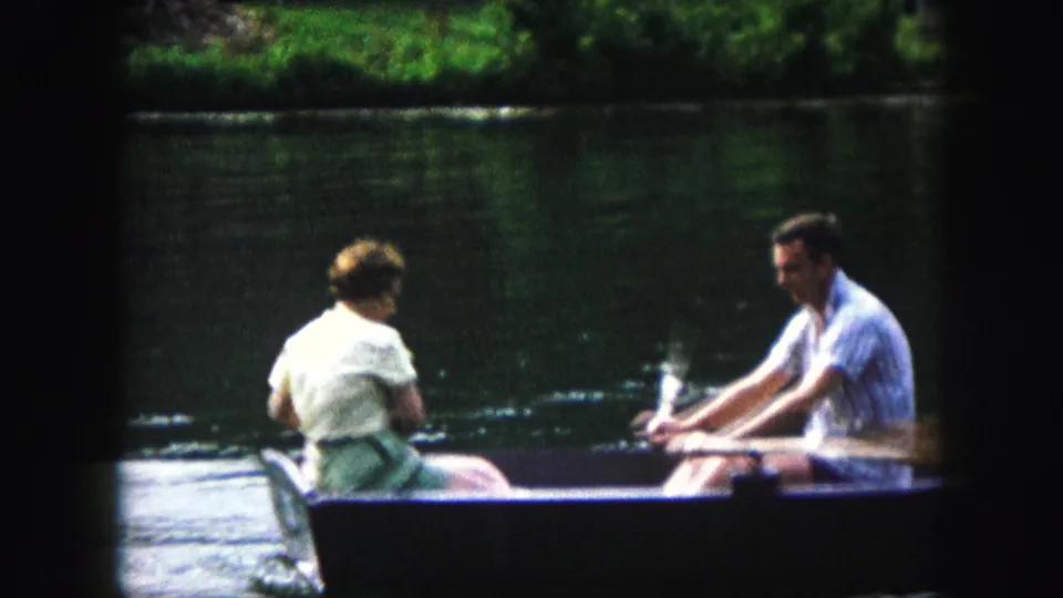 A rear view of the woman and small dog faces the man rowing the boat in soft 8mm color on lake water.