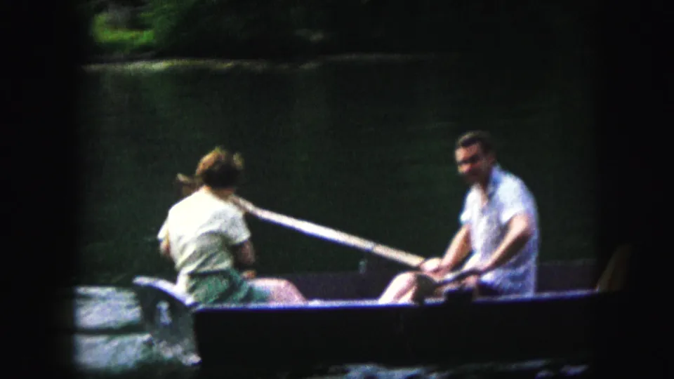 The man smiles toward the camera from the rowboat while the woman and small dog sit opposite him on the lake.