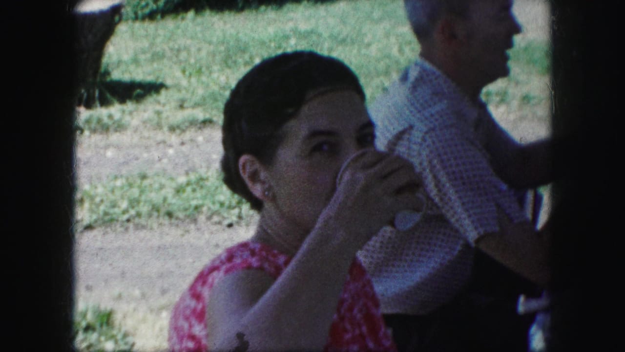 In 1959 Kansas City home-movie footage, a woman drinks from a cup while an older man sits nearby under a picnic shelter.
