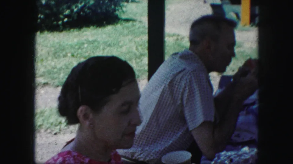A woman in a red patterned dress sits near a cup while an older man eats at a picnic table in 1959 Kansas City footage.