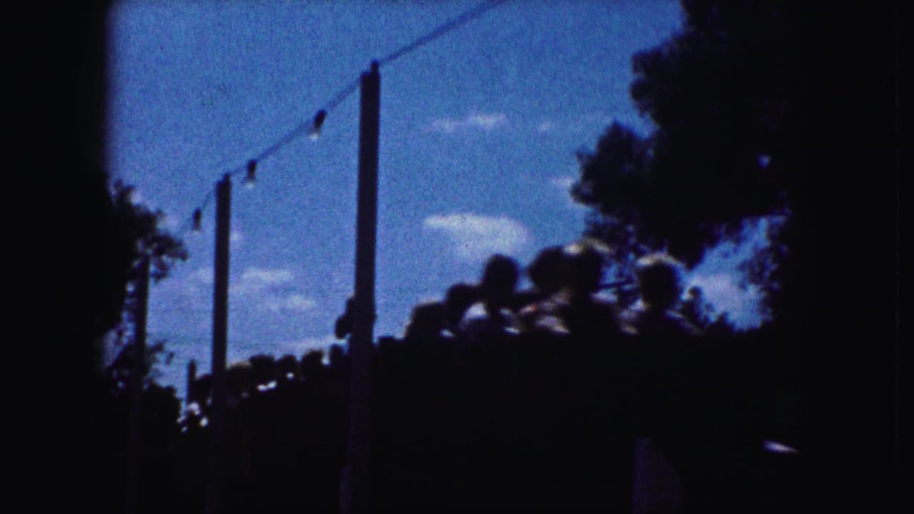 Kansas City 1959 home-movie frame showing silhouetted roller coaster riders under string lights and trees against blue sky.