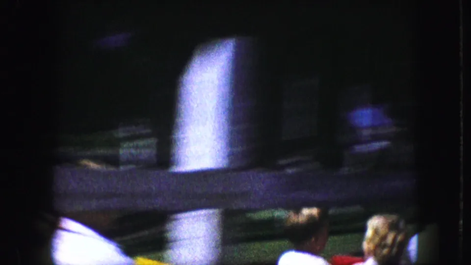 Motion-blurred 1959 coaster footage with dark track structure, a pale support post and riders partly visible at the lower edge.