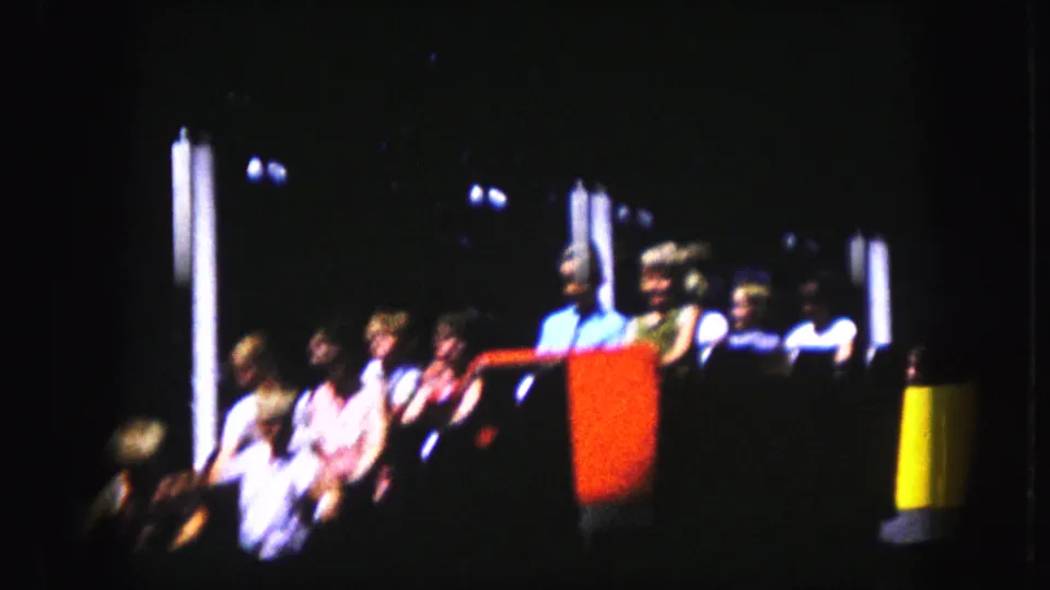1959 Kansas City home-movie frame showing riders seated in a wooden roller coaster car with red and yellow painted sections.
