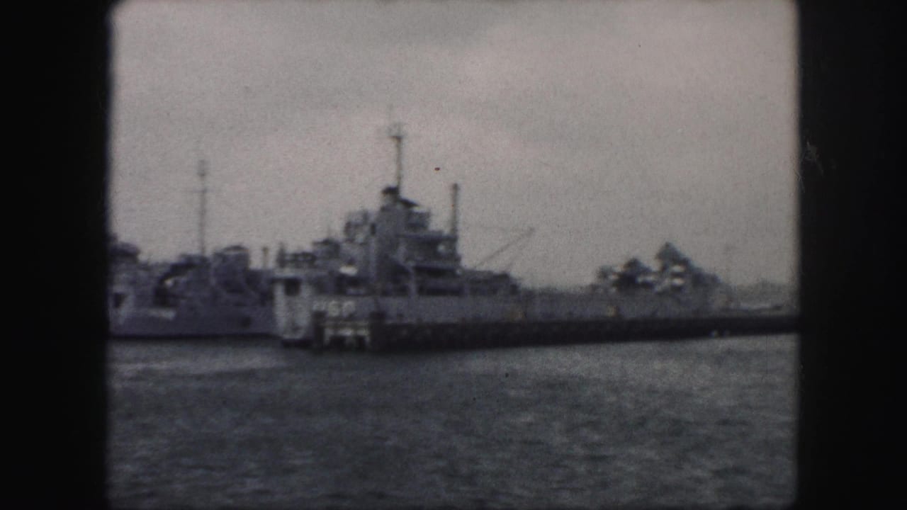 1959 8mm frame of docked military-style ships seen across water, with masts and superstructures silhouetted under overcast sky.