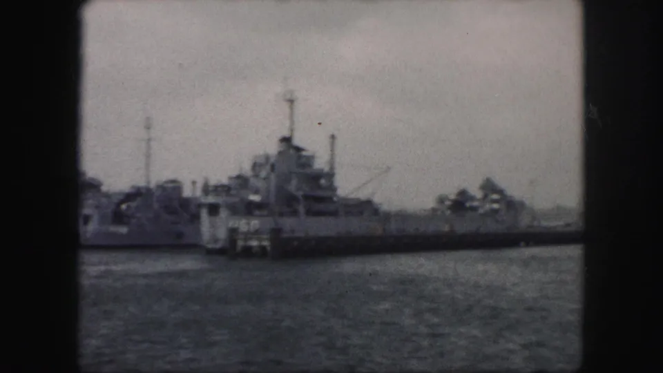 1959 8mm frame of docked military-style ships seen across water, with masts and superstructures silhouetted under overcast sky.