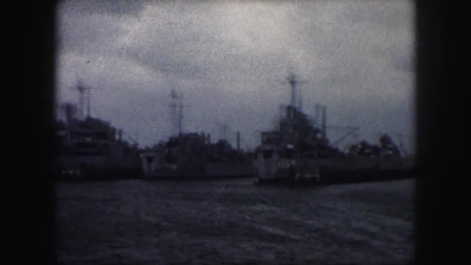 Soft 8mm frame showing several docked military-style vessels, their masts and deck structures visible beyond rippled water.