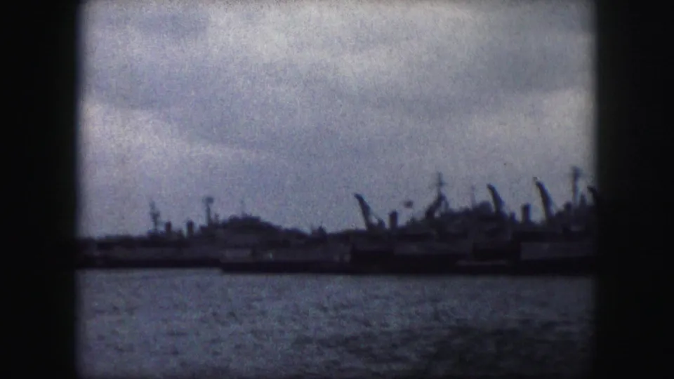 A soft, grainy 1959 home-movie frame shows a low row of docked vessels across water under a pale cloudy sky.