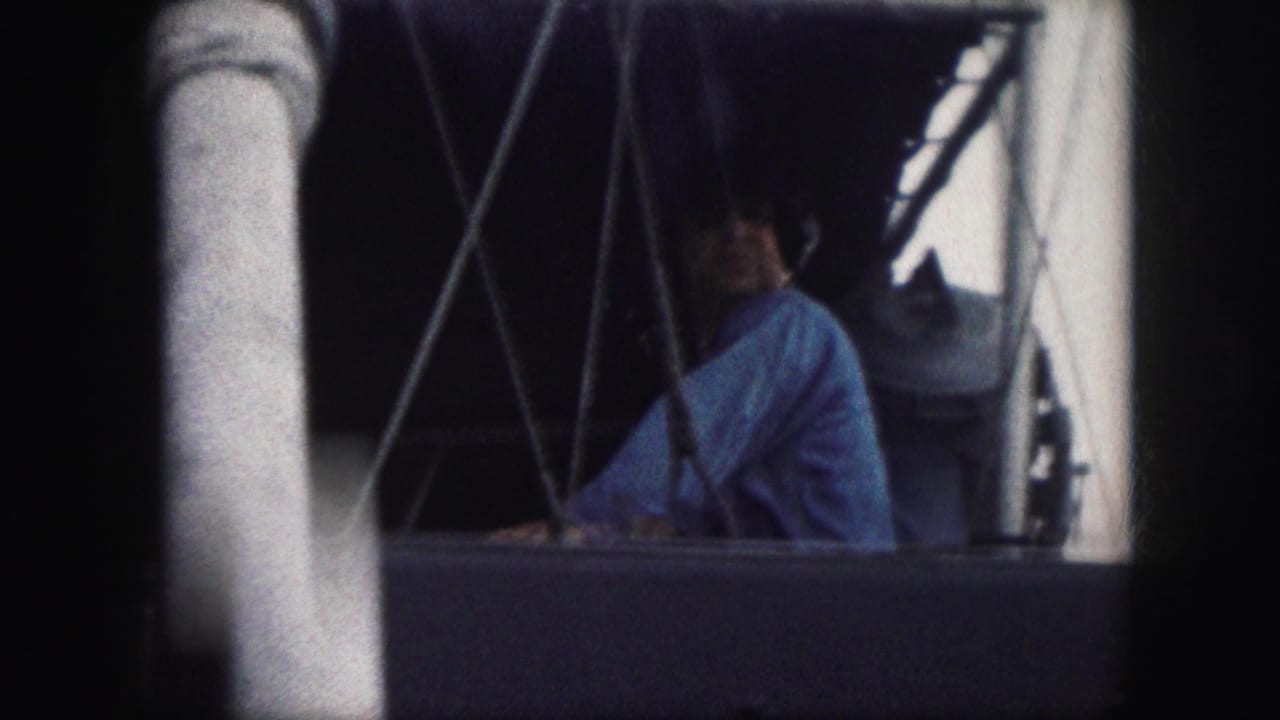 Man in a blue shirt seen through boat framing beside railings and rigging.