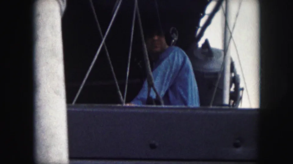 Adult man on a boat in a blue shirt, partially framed by railings, rigging, and dark 8mm borders in the supplied 1959 clip.