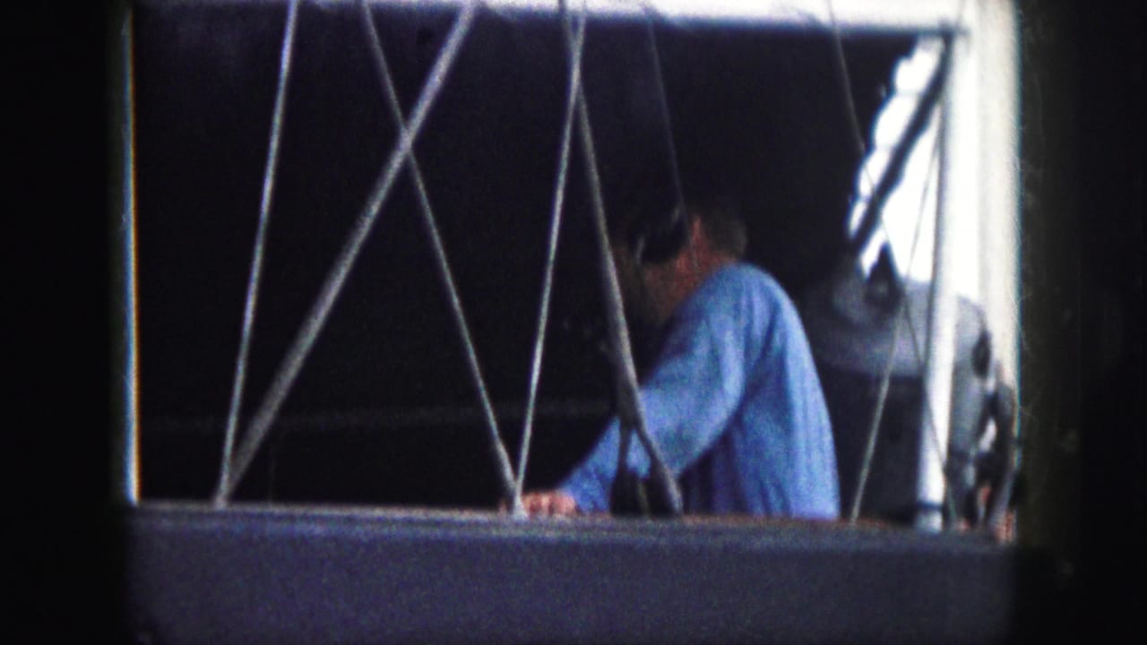 1959 8mm frame from Kansas City metadata showing a man in a blue shirt and headphones behind cross-bracing inside a gondola-like opening.