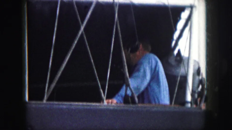 1959 8mm frame from Kansas City metadata showing a man in a blue shirt and headphones behind cross-bracing inside a gondola-like opening.