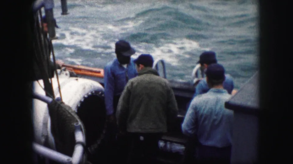 A handheld 8mm frame shows men clustered at the boat rail, with one man partly turned away and another near the ladder.