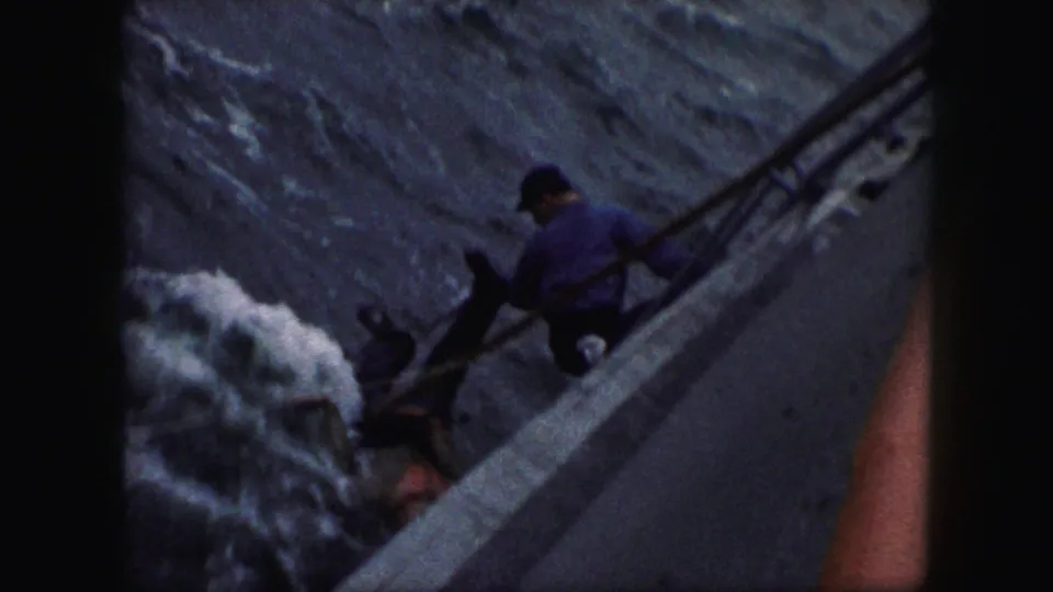 1959 8mm home-movie frame showing a man on a boat reaching down toward a person in rough water, with dark film borders visible.