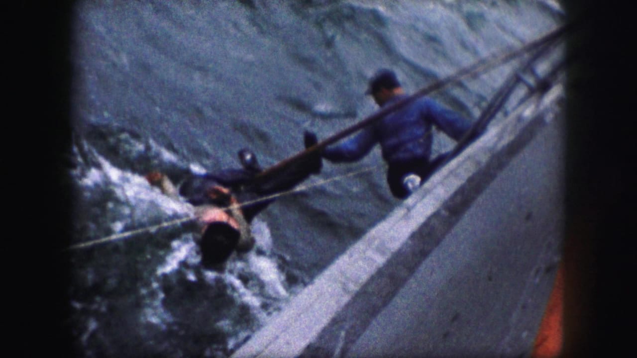 Person in the water is pulled alongside a boat while a man holds the rail above.