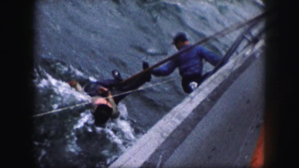 A rough 8mm frame shows a man in the water near the side of a boat as another man in blue braces near the rail.