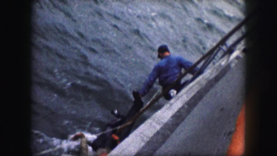 A man in a blue shirt and cap leans from the boat rail toward a partly submerged person, with grainy 8mm borders framing the scene.