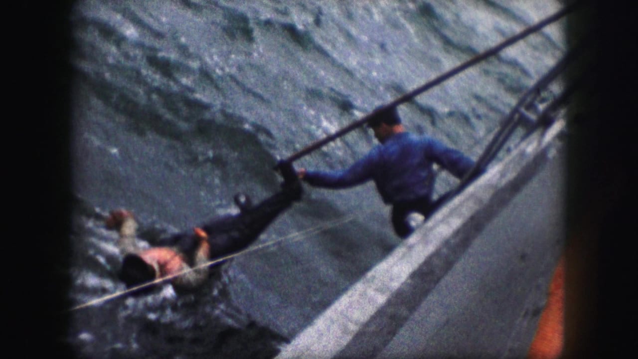 1959 8mm frame showing a person in a blue shirt using a long pole from a vessel rail as another person floats in rough water.